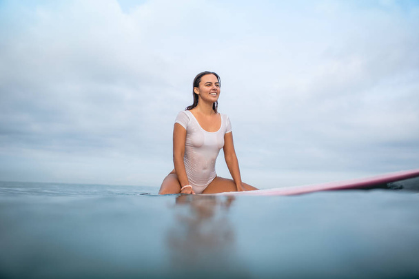 smiling attractive woman in white wet swimsuit sitting on surfboard in ocean during summer vacation - 写真・画像