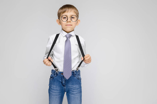 A charming boy in a white shirt, suspenders, a tie and light jeans stands on a gray background. the boy in glasses pulled the suspenders - Photo, Image