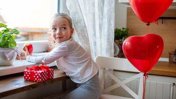 Cute preschooler girl celebrating 6th birthday. Girl with cheeky smile eating her birthday cupcake in the kitchen, surrounded by balloons. - Fotografie, Obrázek