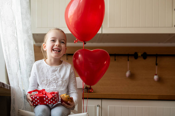 Cute preschooler girl celebrating 6th birthday. Girl holding her birthday cupcake and beautifully wrapped present, sitting in kitchen surrounded by balloons. - Фото, изображение