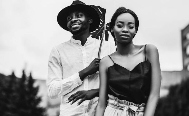 Mooie Afrikaanse jonggehuwden glimlachend buitenshuis poseren met monstera blad, zwart-wit portret - Foto, afbeelding