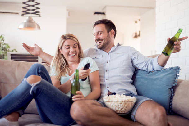 Young couple relaxing at home, sitting on couch, watching movie and eating popcorn - Photo, image