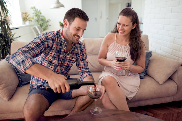 Caucásico pareja beber vino mientras está sentado en el sofá en la sala de estar
 - Foto, Imagen