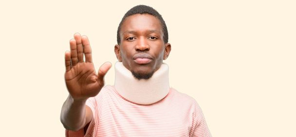 Hombre negro africano lesionado con corsé en el cuello molesto con mala actitud haciendo señal de stop con la mano, diciendo que no, expresando seguridad, defensa o restricción, tal vez empujando
 - Foto, imagen