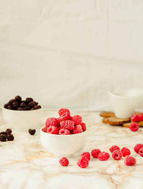 Raw raspberries and wild black berries in white cups. Light marb - Valokuva, kuva
