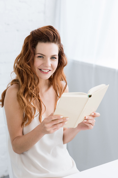 happy young woman reading book at home and looking at camera - Fotoğraf, Görsel
