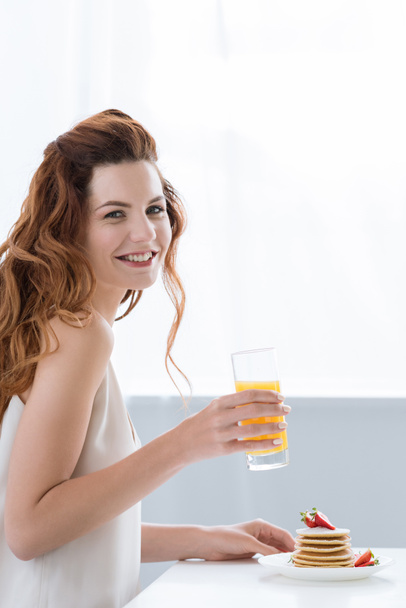 smiling young woman with orange juice and pancakes with strawberry for breakfast looking at camera - Foto, afbeelding
