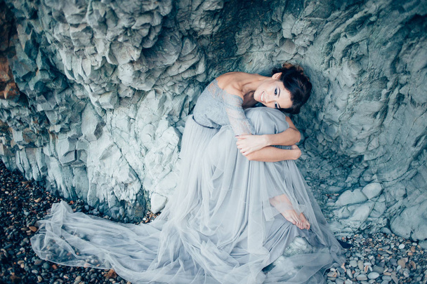 Very beautiful brunette girl in a long gray gown of lace and tulle, the veil sits with his head bowed and hugging his knees with smile on her face, near the rocks - Φωτογραφία, εικόνα
