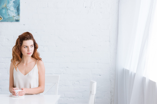 thoughtful young woman with cup of coffee in front of white brick wall looking at window - Photo, Image