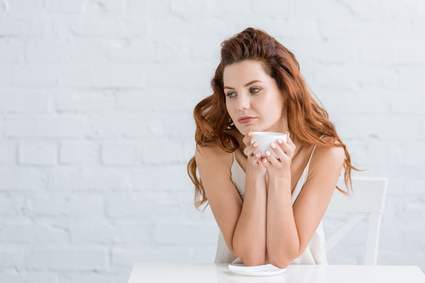 Nachdenkliche junge Frau mit Tasse Kaffee vor weißer Backsteinwand - Foto, Bild