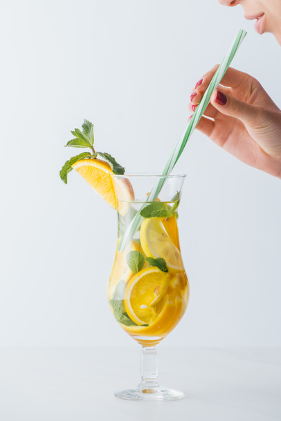 partial view of woman holding straw and summer cold cocktail with mint, lemon and orange pieces isolated on white - Foto, imagen