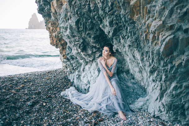 Beautiful brunette girl in a long grey dress with a veil sitting by the sea, near the cliff, - Foto, afbeelding