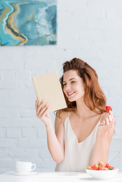 happy young woman with coffee and strawberry reading book at home - 写真・画像