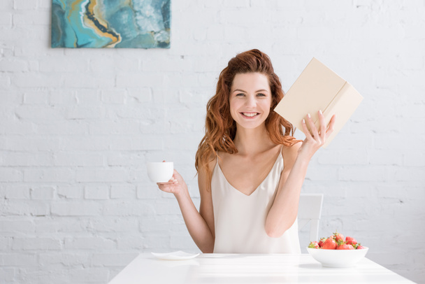 glückliche junge Frau mit Buch und Tasse Kaffee, die zu Hause in die Kamera schaut - Foto, Bild