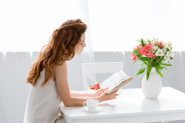 mujer joven enfocada leyendo libro mientras está sentada en la mesa con taza de café y flores en jarrón
 - Foto, imagen