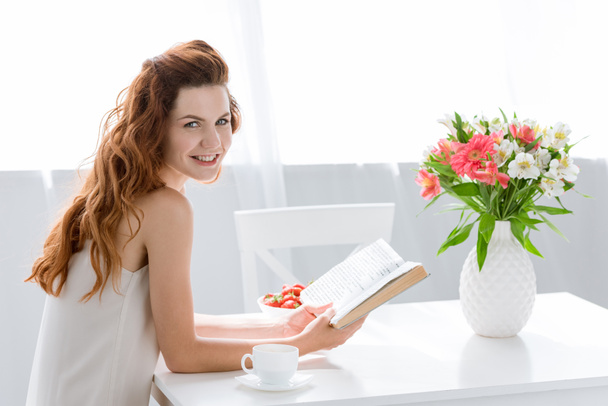 happy young woman reading book while sitting at table with coffee cup and flowers in vase - Foto, Imagen