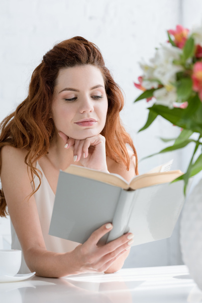 ελκυστική νεαρή γυναίκα διαβάζοντας το βιβλίο ενώ κάθεται στο τραπέζι με φλιτζάνι καφέ και λουλούδια σε βάζο - Φωτογραφία, εικόνα