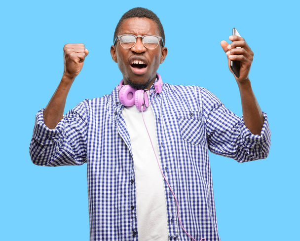 Αφρικανική μαύρο άντρα φοιτητή με smartphone χαρούμενος και συγκινημένος γιορτάζουμε νίκη εκφράζοντας την μεγάλη επιτυχία, δύναμη, ενέργεια και θετικά συναισθήματα. Γιορτάζει χαρούμενη νέα δουλειά - Φωτογραφία, εικόνα