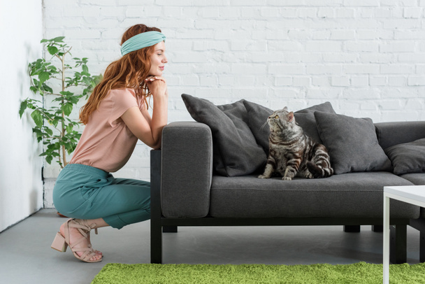 усміхнена молода жінка дивиться на свого таббі-кішку, сидячи на дивані вдома
 - Фото, зображення