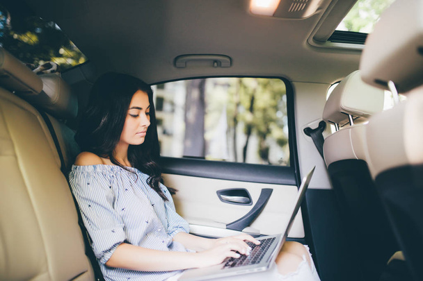Όμορφη γυναίκα είναι χρησιμοποιώντας ένα φορητό υπολογιστή και χαμογελώντας, ενώ κάθονται στο πίσω κάθισμα στο αυτοκίνητο - Φωτογραφία, εικόνα