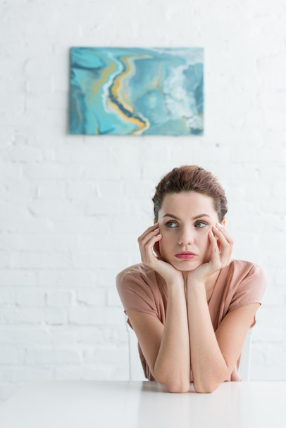 thoughtful young woman sitting at table in front of white brick wall with picture - Photo, Image
