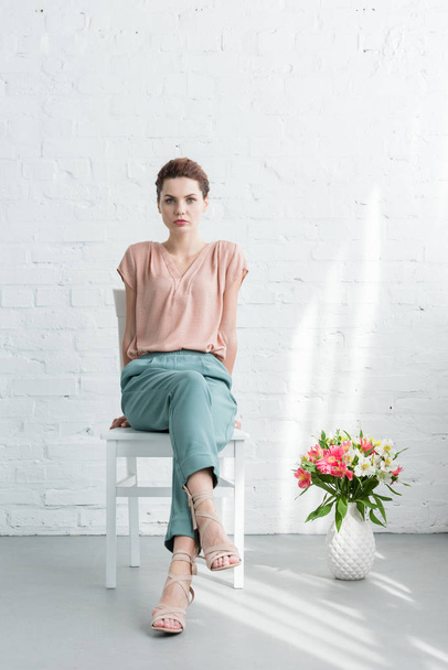 jolie jeune femme assise sur une chaise avec des fleurs sur le sol devant un mur de briques blanches
 - Photo, image