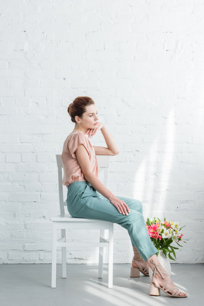 στοχαστικός νεαρή γυναίκα που κάθεται σε καρέκλα με λουλούδια στο πάτωμα μπροστά από το λευκό τοίχο - Φωτογραφία, εικόνα