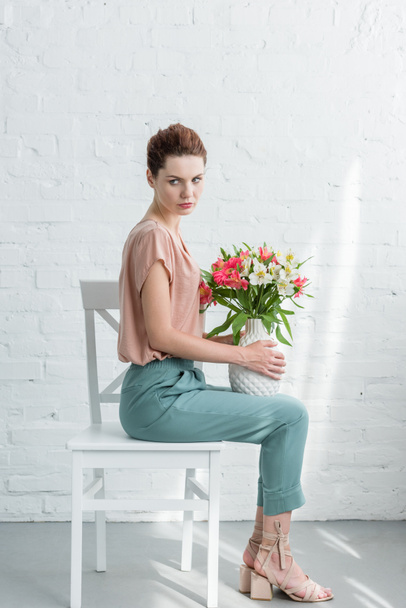 Seitenansicht der schönen jungen Frau, die eine Vase mit Blumen hält, während sie auf einem Stuhl vor einer weißen Backsteinwand sitzt - Foto, Bild