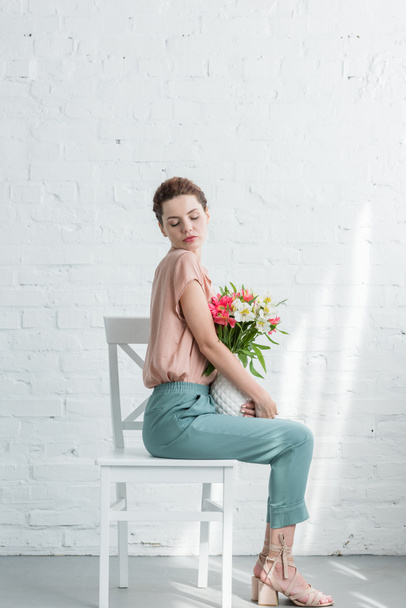 schöne junge Frau mit frischen Blumen in Vase vor weißer Ziegelwand - Foto, Bild