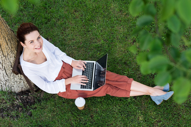 top view of beautiful young woman working with laptop while sitting on grass with paper cup of coffee - Foto, Imagen