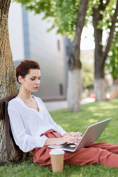 hermosa joven freelancer trabajando con el ordenador portátil mientras se apoya de nuevo en el tronco del árbol en el parque
 - Foto, imagen