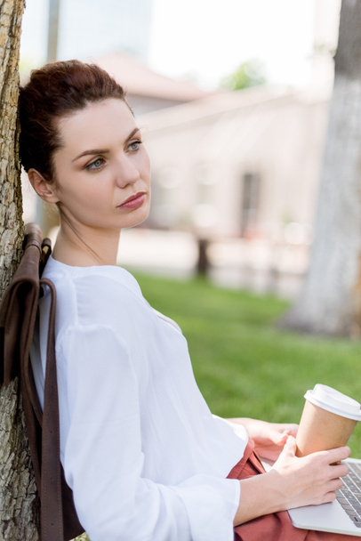 Nachdenkliche junge Frau mit Laptop und Pappbecher Kaffee lehnt sich an Baumstamm im Park zurück und schaut weg - Foto, Bild