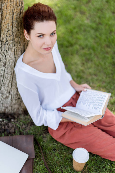 vista de ángulo alto de la joven feliz con libro apoyado en el tronco del árbol en el parque y mirando a la cámara
 - Foto, Imagen