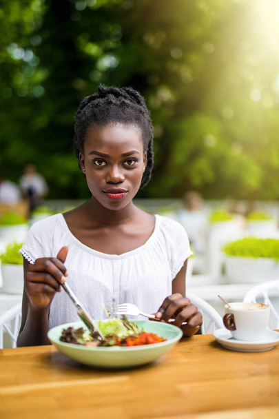 Mujer afroamericana joven sentada en la cafetería al aire libre comer ensalada fresca al aire libre
 - Foto, imagen