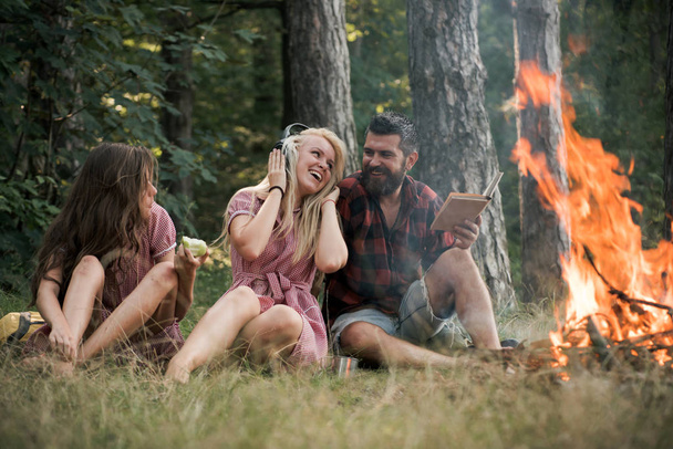 Friends relax at bonfire flame with sparks in vintage style. People camping at fire in forest. Women and bearded man at campfire. Eating food, reading book and entertainment. Summer vacation concept - Fotó, kép