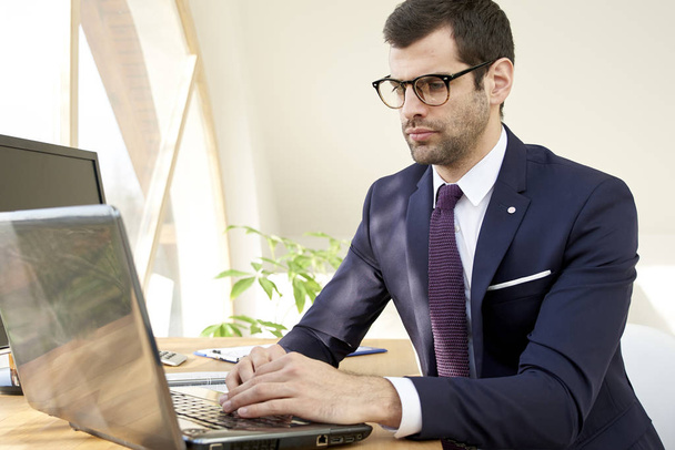 Portrait of young businessman in suit and glasses using laptop while sitting at office desk and working on new project.  - Fotoğraf, Görsel