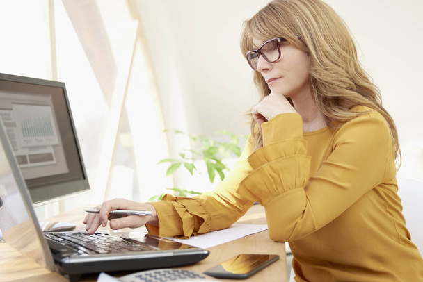 Middle aged casual investment advisor businesswoman working on laptop while sitting at office desk.  - Foto, imagen