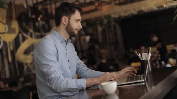 Man with a laptop at a table in a restaurant or bar, man working on laptop in cafe,Young hipster man having a coffee break at the bar, he is holding a cup and connecting with a laptop, businessman - Кадры, видео