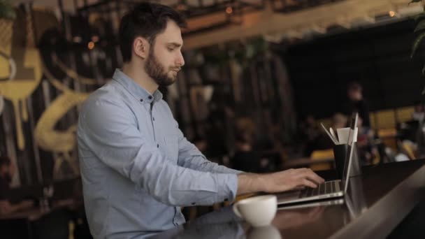 Мужчина с ноутбуком за столом в ресторане или баре, мужчина, работающий на ноутбуке в кафе, молодой хипстер с кофе-брейком в баре, он держит чашку и соединяется с ноутбуком, бизнесмен
 - Кадры, видео