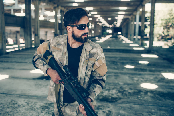 Serious and handsome man in glasses is standing in hangar and holding paintball gun in hands. He is looking straight forward. Man wears special uniform. - Φωτογραφία, εικόνα