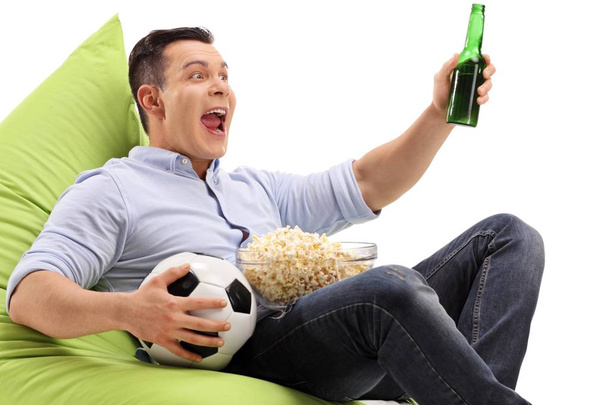 Young man with a bottle of beer and a bowl of popcorn sitting on a beanbag and watching soccer on television isolated on white background - Фото, зображення