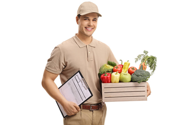 Delivery man holding a crate filled with groceries and clipboard isolated on white background - Fotó, kép