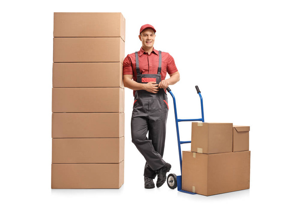 Full length portrait of a mover with a hand truck leaning on a stack of boxes and smiling isolated on white background - Fotografie, Obrázek