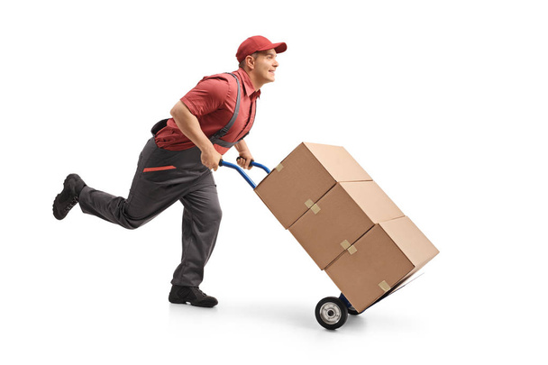 Full length profile shot of a mover running and pushing a hand truck loaded with boxes isolated on white background - Zdjęcie, obraz