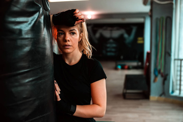 Beautiful exhausted woman leaning on punching bag at gym  - Photo, Image