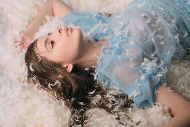 Lady in transparent blue nightie lay on bed in heap of feathers. Woman with long hair in tender pajama relaxing. Girl on mysterious face lay on bed covered with feathers and fluff. Perfume concept. - Valokuva, kuva