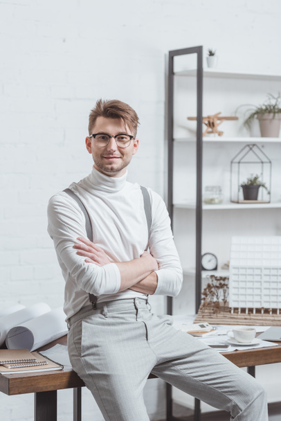 portrait of smiling architect in eyeglasses with arms crossed leaning on workplace in office - Photo, Image