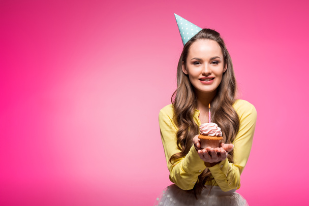 attractive woman with party hat holding cupcake with candle isolated on pink - Photo, Image