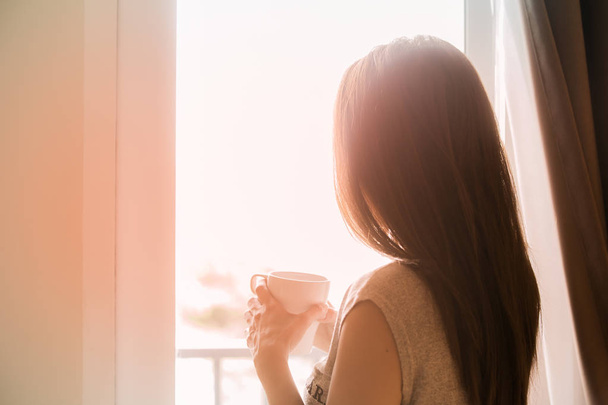 Donna in piedi che tiene la tazza di caffè guardando fuori dalla finestra
. - Foto, immagini