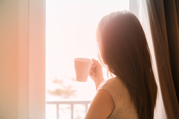 Standing woman holding coffee cup looking out the window. - 写真・画像